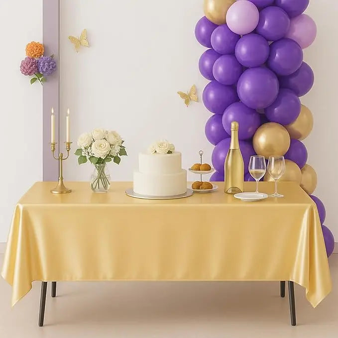 A gold satin tablecloth is set for an elegant event with a white cake, flowers, and a bottle of champagne.