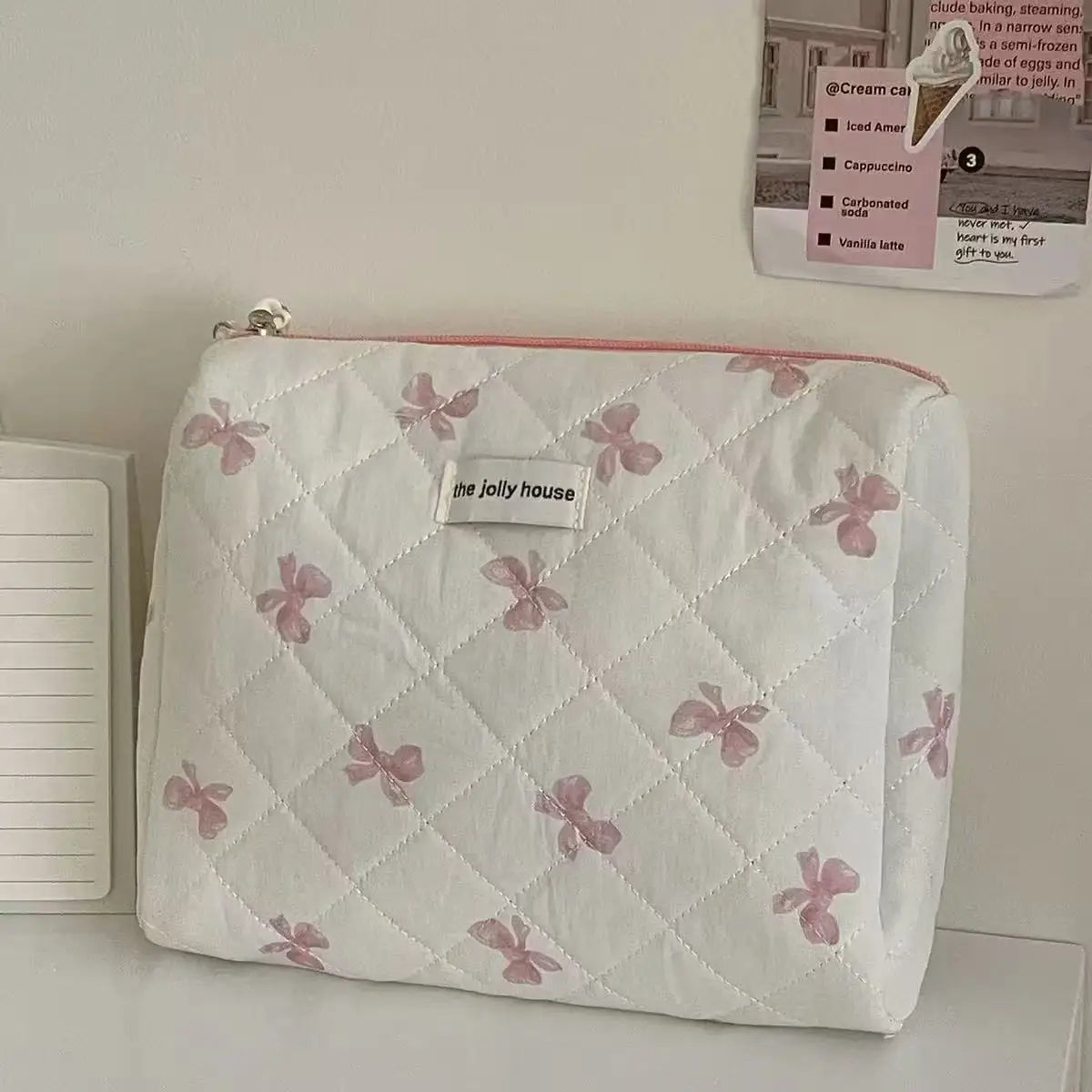 A white cosmetic bag with a pink bow print and quilted texture resting on a white surface.