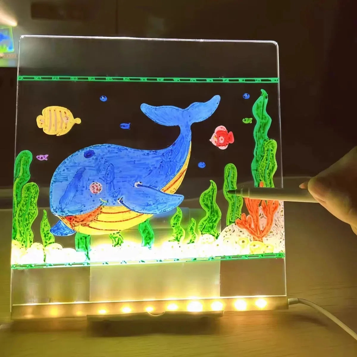 A glowing LED drawing board displays a blue whale and various green and orange colored pens on a dark background.