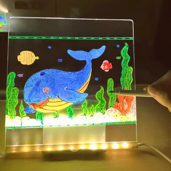 A glowing LED drawing board displays a blue whale and various green and orange colored pens on a dark background.