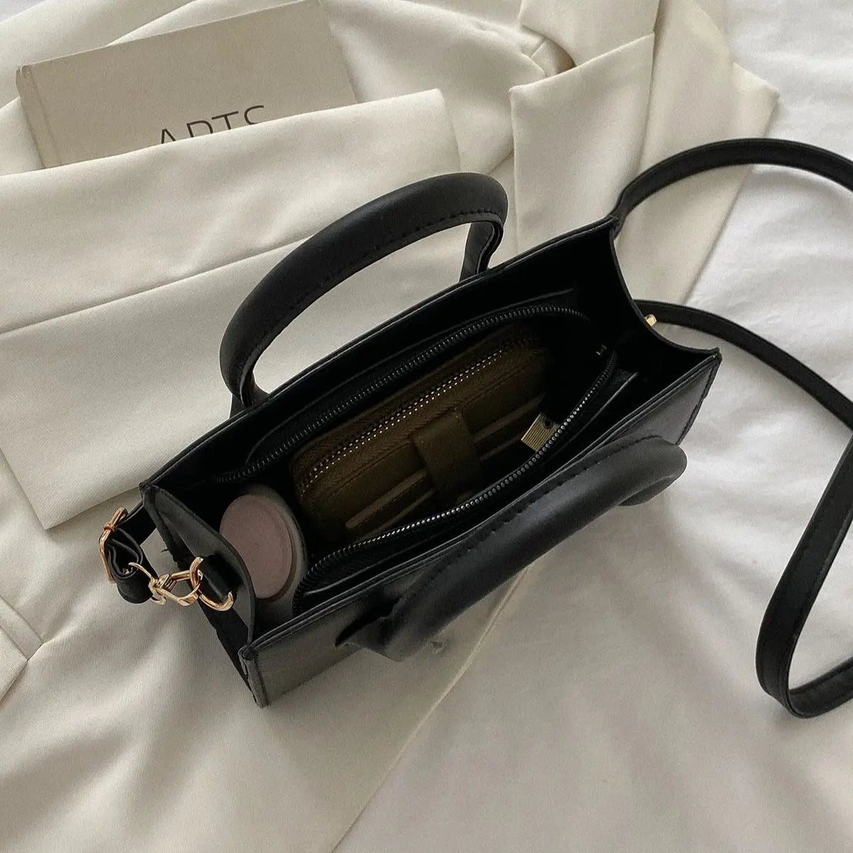 A black PU leather crossbody tote bag with a gold chain detail resting on a white fabric background.