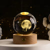 A laser engraved crystal ball night light with a golden glow resting on a wooden base.
