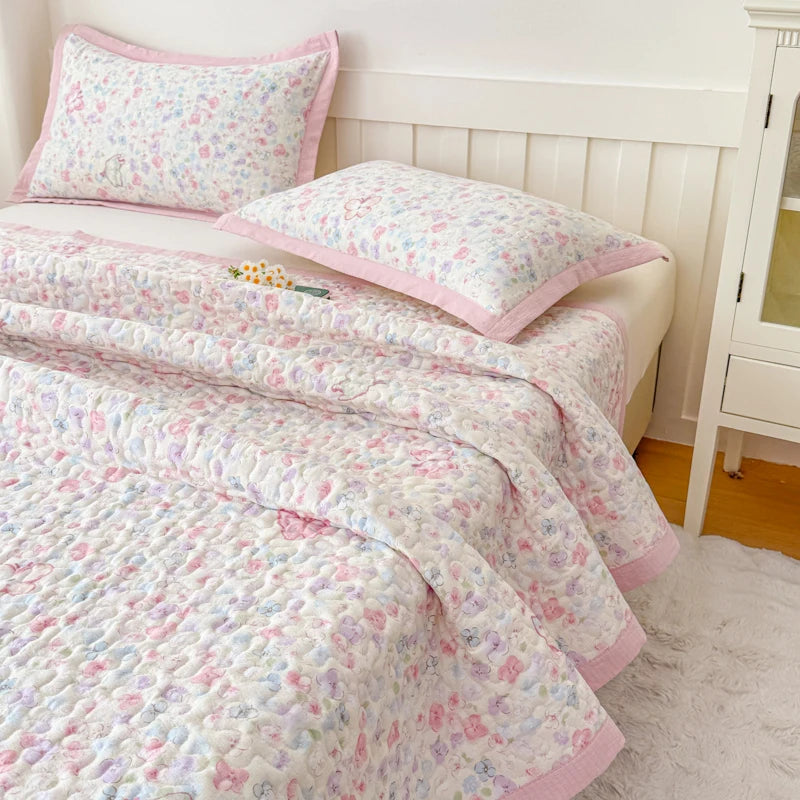 A pink and white floral quilted bedspread with a pink trim is displayed on a bed, next to a white wooden cabinet.