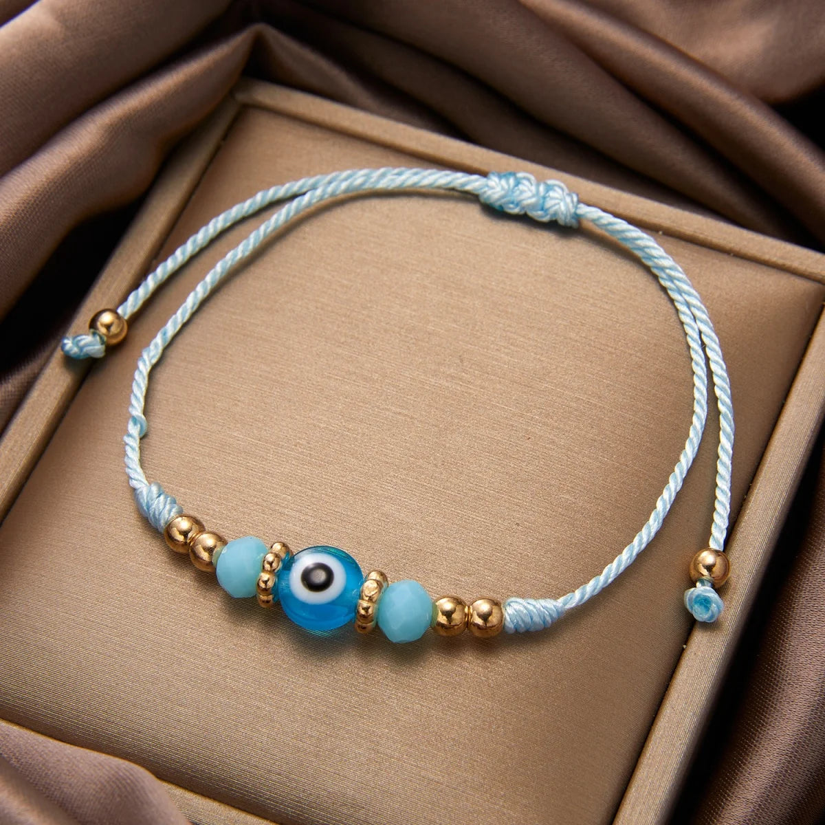 A light blue crystal bead bracelet with an evil eye charm and gold accents resting on a beige fabric background.