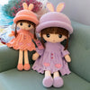 A pair of soft plush dolls resembling rabbits, one in peach and one in lavender, sitting on a green sofa.