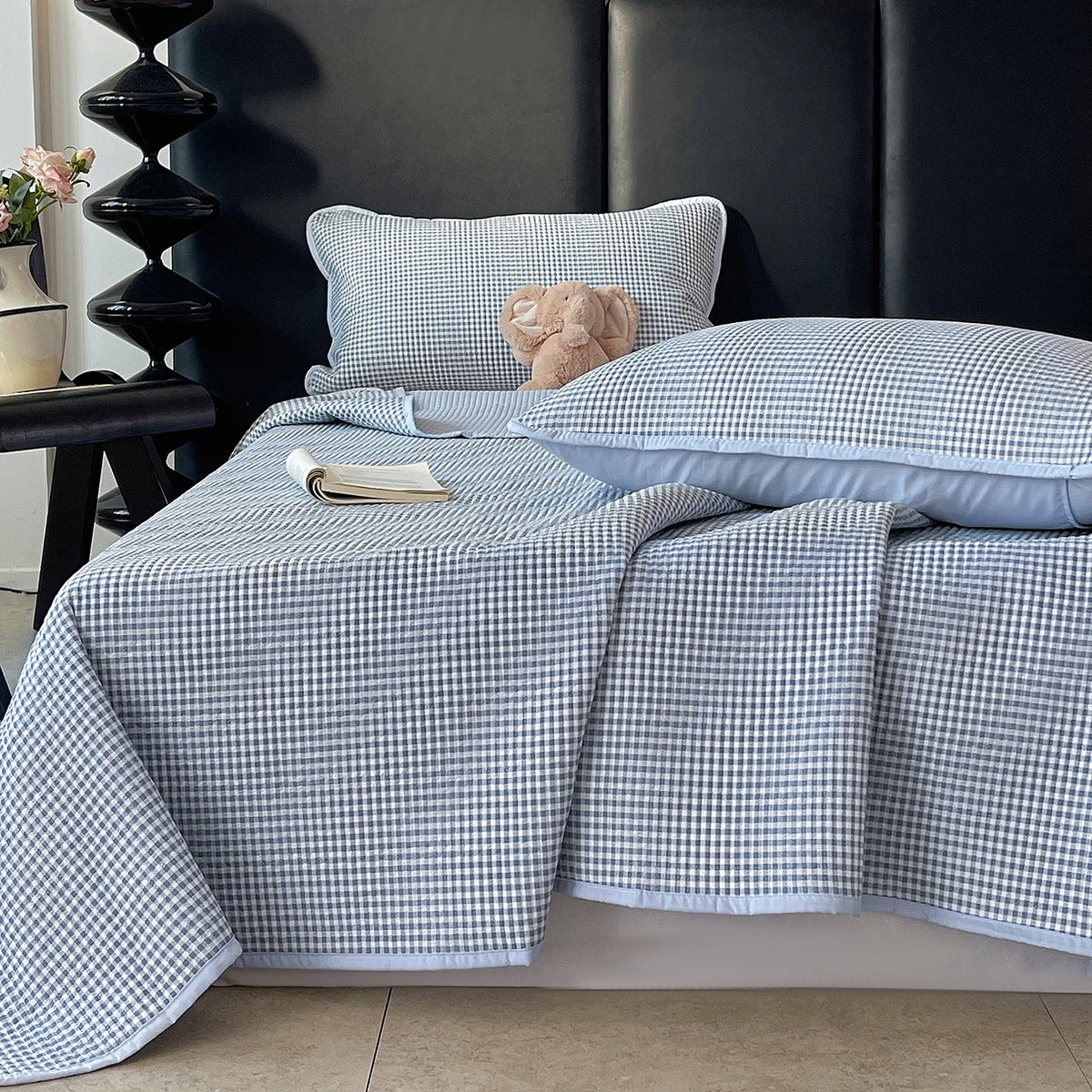 A light blue and white checkered quilted bedspread with a fluffy teddy bear pillow on a dark headboard.