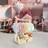 A wooden storage crate with a handle is filled with baby essentials and toys, set in a pink decorative scene.
