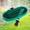 A coil of green flat soaker hose with a spray nozzle lying on a green lawn amidst garden foliage.
