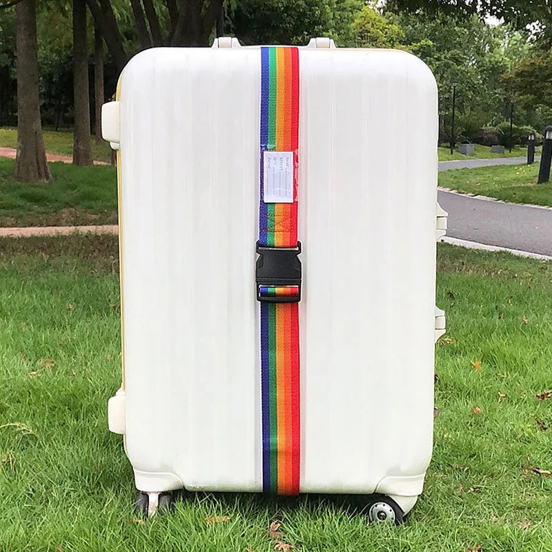 A white suitcase with colorful adjustable straps is placed on green grass next to a paved path.