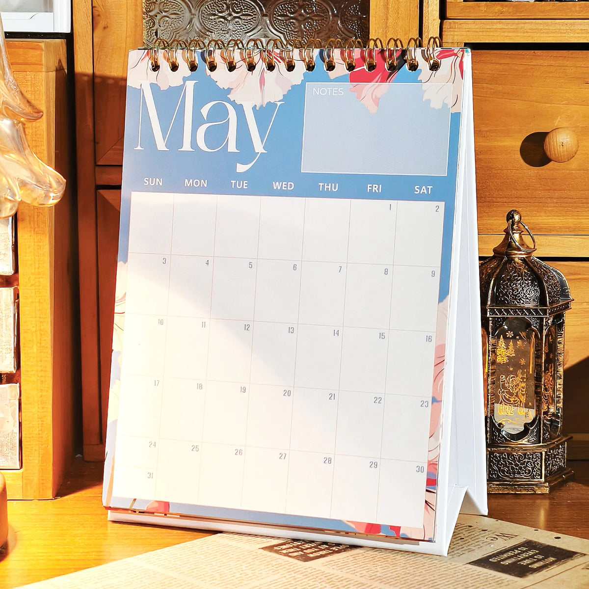A vertical desk calendar for May featuring a monthly planner layout with floral accents, placed on a wooden surface.