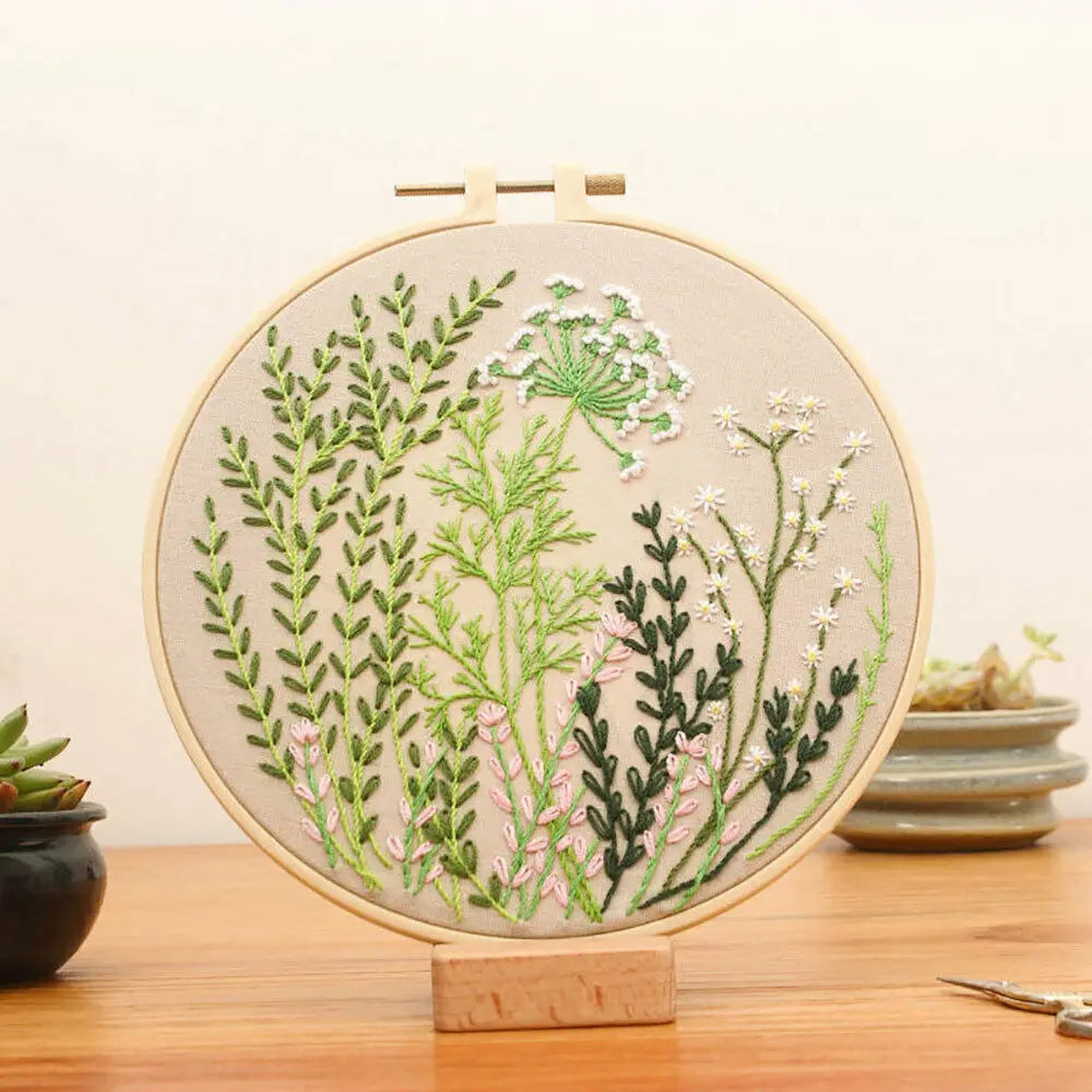 A circular embroidery hoop featuring floral cross stitch designs on a beige fabric, displayed on a wooden stand.