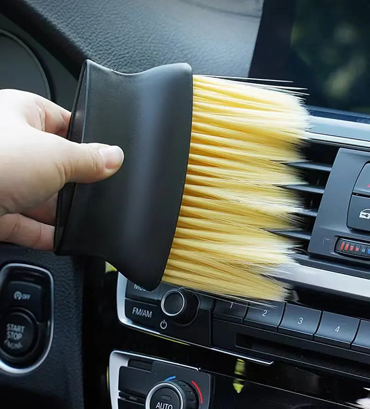 A hand holds a black detailing brush set with soft yellow bristles, positioned near a car interior panel.
