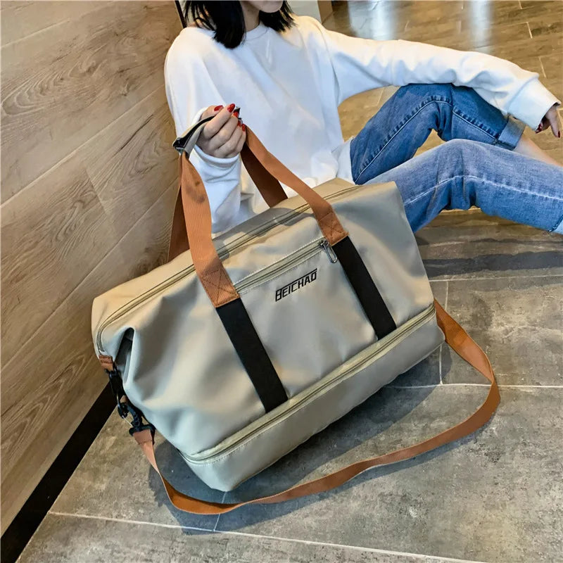 A beige travel duffle bag with brown leather accents is placed on a floor next to a person wearing a white top and blue jeans