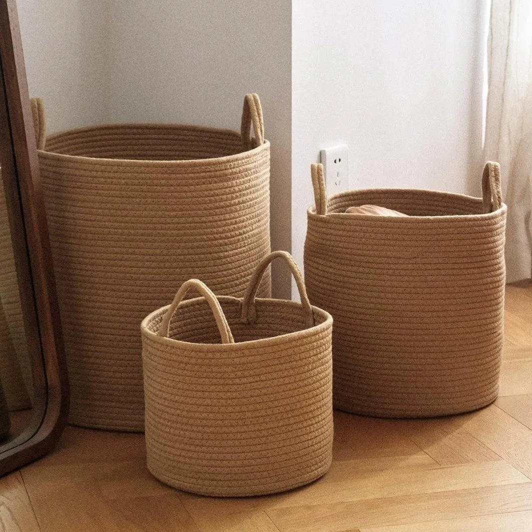 Three natural woven storage baskets of varying sizes made from cotton rope with handles on a wooden floor.