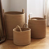 Three natural woven storage baskets of varying sizes made from cotton rope with handles on a wooden floor.