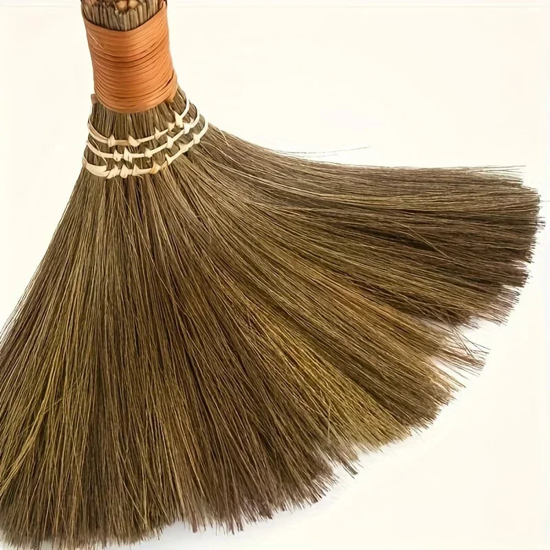 A handmade natural grass broom with a woven handle and light brown bristles is shown in a close-up view.