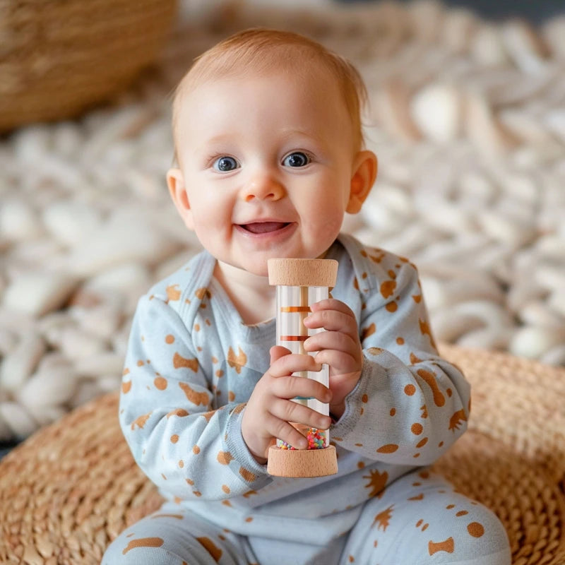 A smiling baby holding a wooden rain rattle sensory toy, sitting on a woven mat.