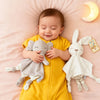 A baby is holding a gray elephant plush toy with ears and a white bunny plush toy on a pink blanket.