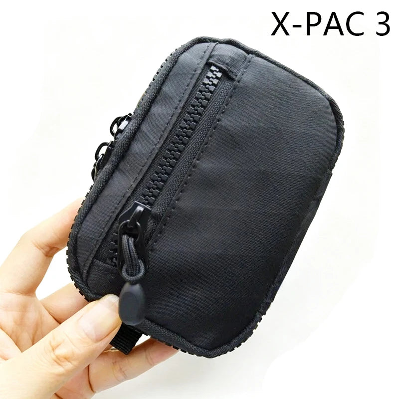 A hand holds a black X-PAC 3 utility pouch featuring a zipper and a toggle closure.