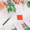 Hands are painting a diamond painting design featuring green and yellow stones on white paper.
