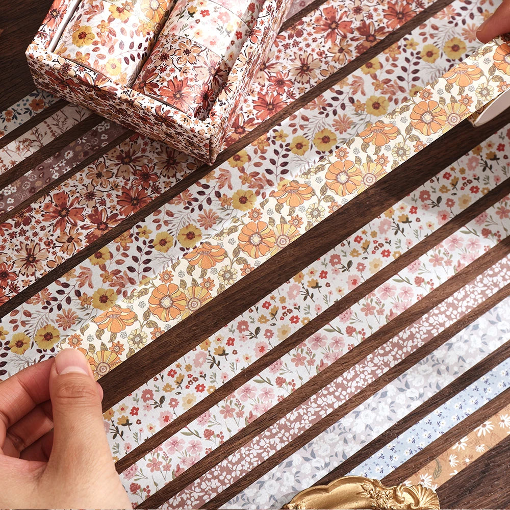 Une main décolle un rouleau de ruban washi floral vintage de sa boîte sur une surface à motifs.