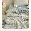 A striped light blue and white quilt is displayed on a bed, with a matching pillow and a small wooden lamp nearby.