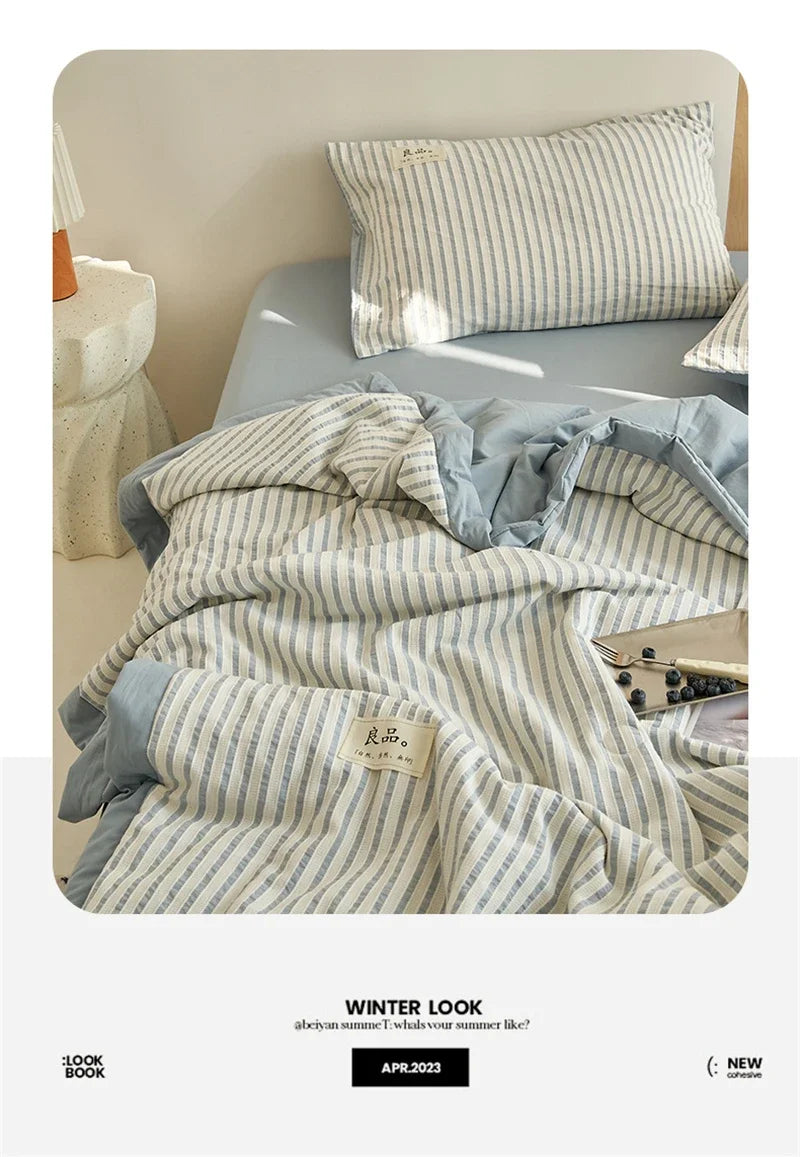 A striped light blue and white quilt is displayed on a bed, with a matching pillow and a small wooden lamp nearby.