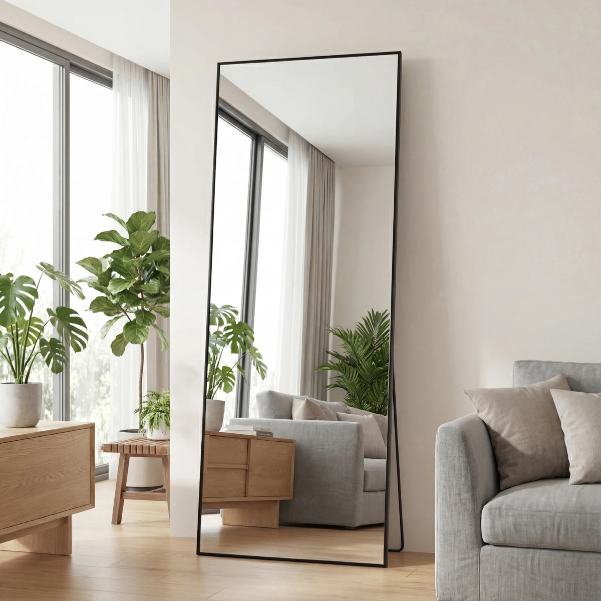 A full length rectangular mirror with a black frame leaning against a white wall in a bright room.