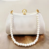 A white satin evening clutch with a gold chain and pearl embellishments displayed on a wooden surface.