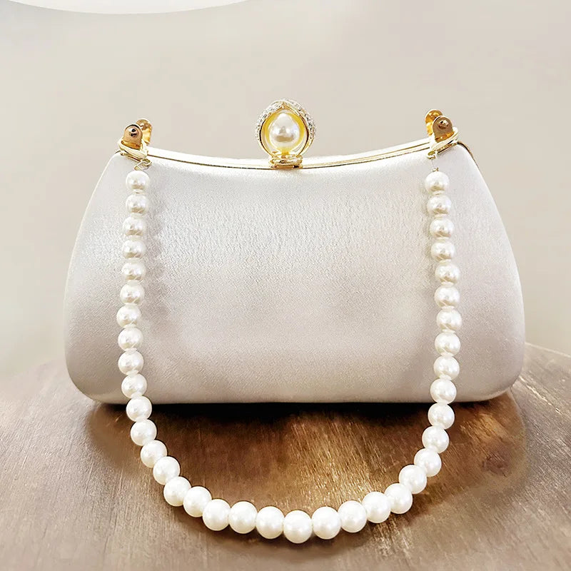 A white satin evening clutch with a gold chain and pearl embellishments displayed on a wooden surface.