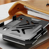 A silver carbon steel tablet stand is shown resting on a light-colored desk next to a brown leather folder.