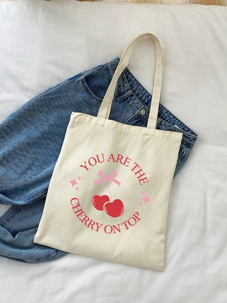 A beige canvas tote bag with pink cherry embroidery that reads You are the cherry on top, resting on a denim shirt.