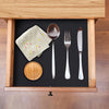 A wooden drawer holds silverware, a napkin, and a wooden plate on a black shelf liner mat.