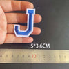 A hand holds a blue embroidered letter J patch, measuring 5 by 3.6 centimeters against a ruler.
