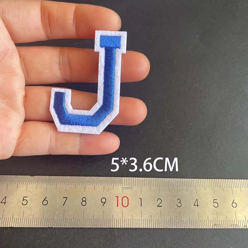 A hand holds a blue embroidered letter J patch, measuring 5 by 3.6 centimeters against a ruler.