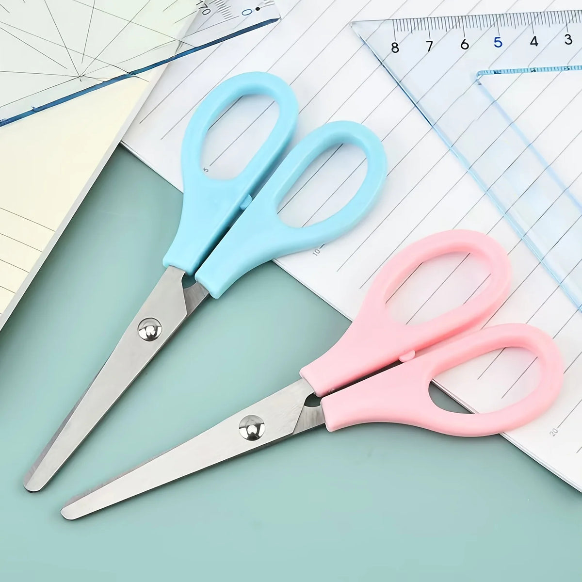 A pair of precision stainless steel scissors in light blue and pink resting on a sheet of paper with a ruler.