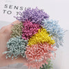 A hand holds a cluster of 400 pearl flower stamens in various shades of purple, pink, and blue for DIY crafts.