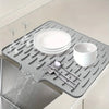 A silicone dish drying mat is shown with dishes and silverware being dried under running water.