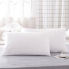 A set of two white microfiber pillowcases resting on a gray surface near a window with sheer curtains.