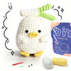 A white crocheted duck with green plant elements, next to yarn and a crochet needle.