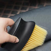 A hand holds a black detailing brush set with yellow soft bristles against a dark gray car interior surface.