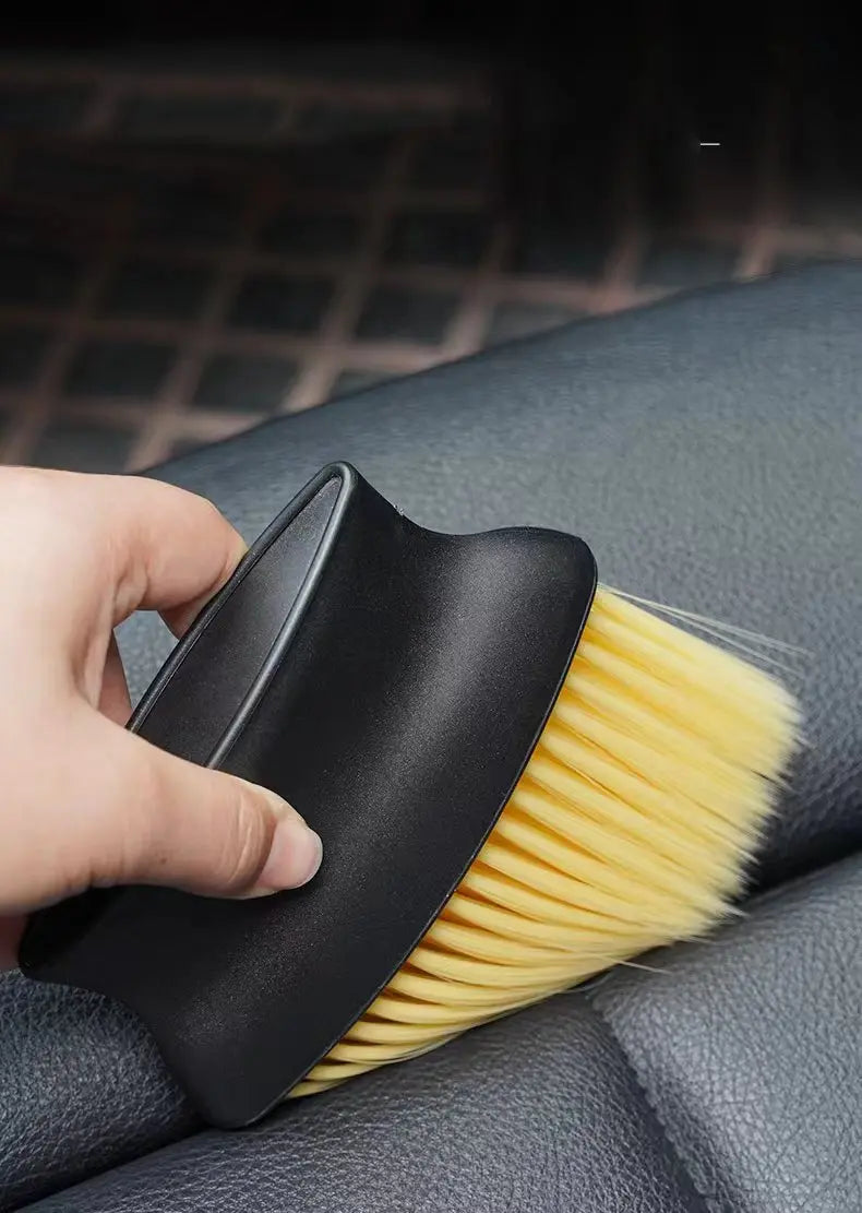 A hand holds a black detailing brush set with yellow soft bristles against a dark gray car interior surface.