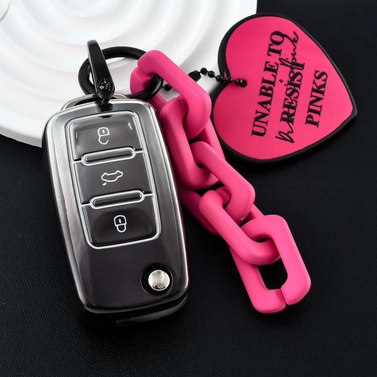 A silver car key fob attached to a bright pink chain, next to a pink heart-shaped tag.