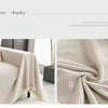 A close-up of an elegant beige herringbone chenille sofa throw with tassel edges draped on a light-colored surface.