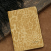 A tan leather passport holder with a leopard print pattern and personalized name embossing rests on a brown surface.