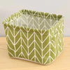 A square storage basket made of green fabric with a white geometric pattern, resting on a wooden surface.