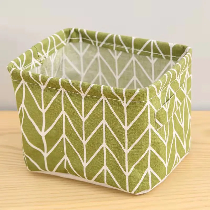 A square storage basket made of green fabric with a white geometric pattern, resting on a wooden surface.