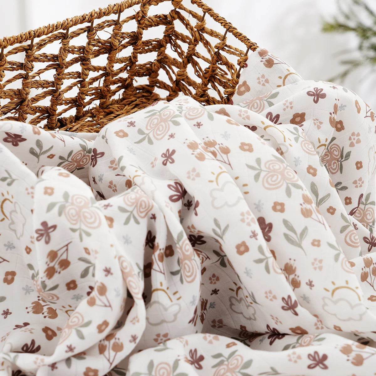 A close-up of a muslin baby swaddle blanket in white fabric with floral patterns, resting on a woven natural fiber mat.