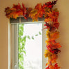 An artificial maple leaf garland in autumn colors hangs on a window frame, decorated with green vines.