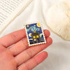 A hand holds a vintage airline ticket enamel brooch set featuring a police box design on a square piece of enamel.